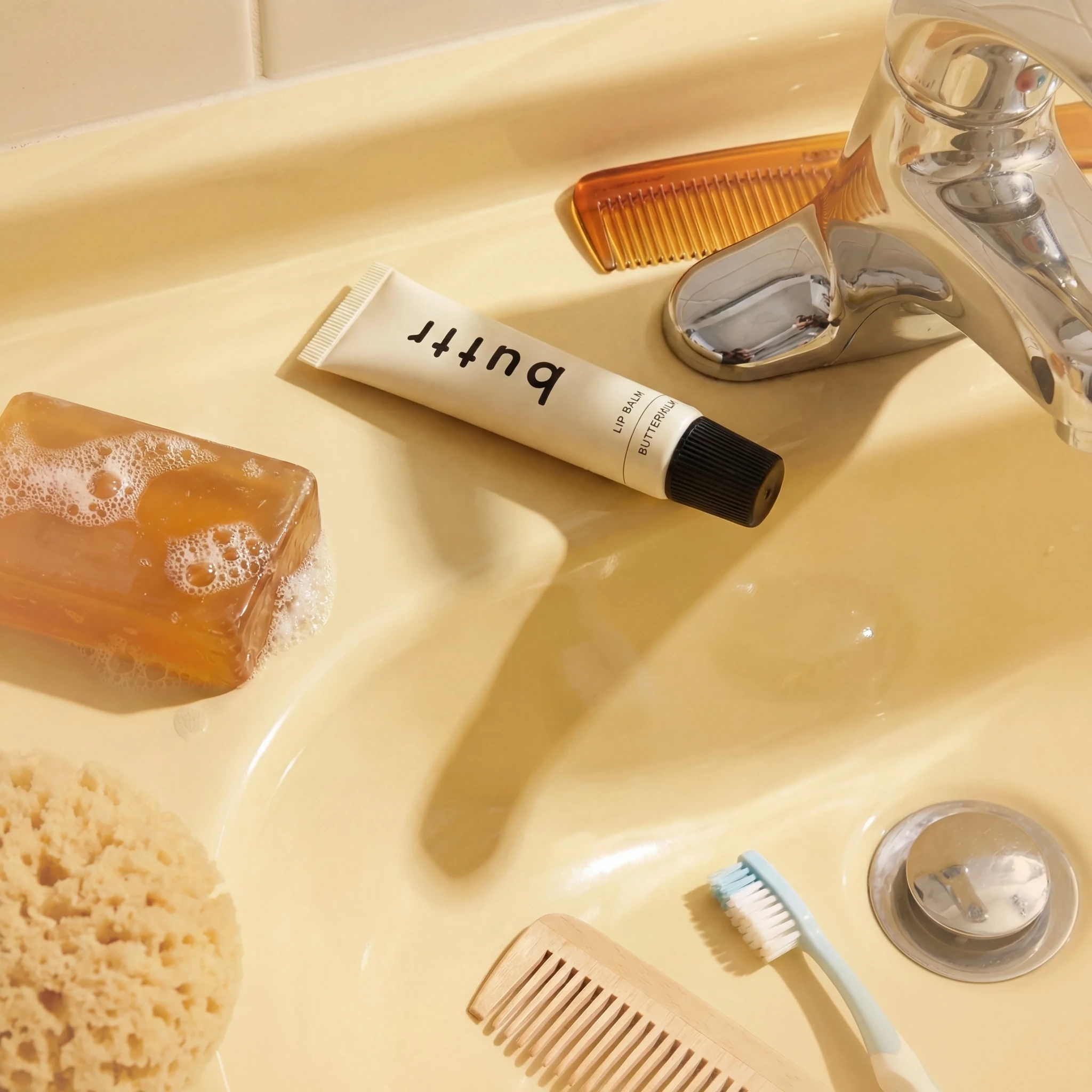 Lip balm styled beside soap, combs and a toothbrush on a yellow bathroom sink