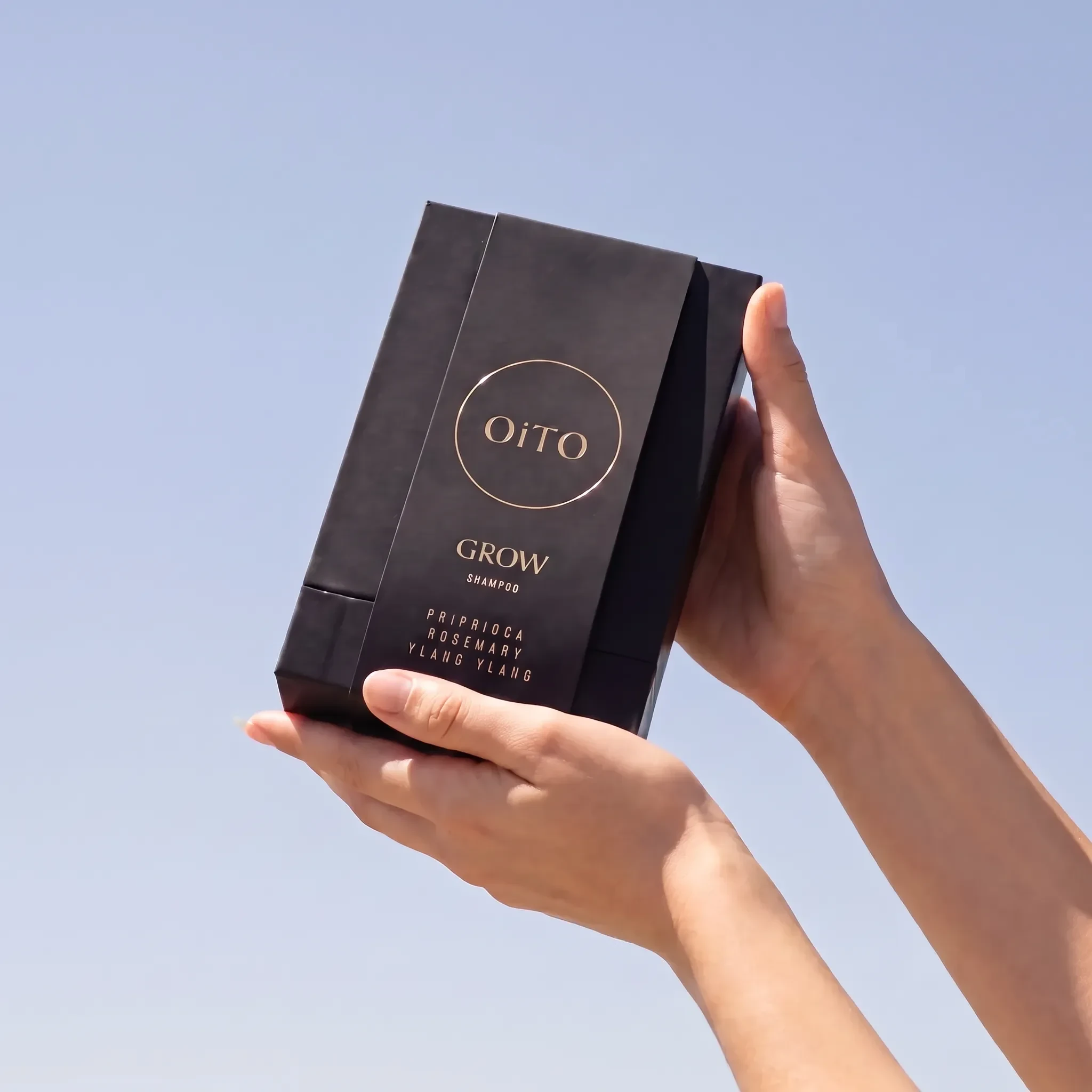 Hands holding an OiTO Grow Shampoo box against a pale blue background