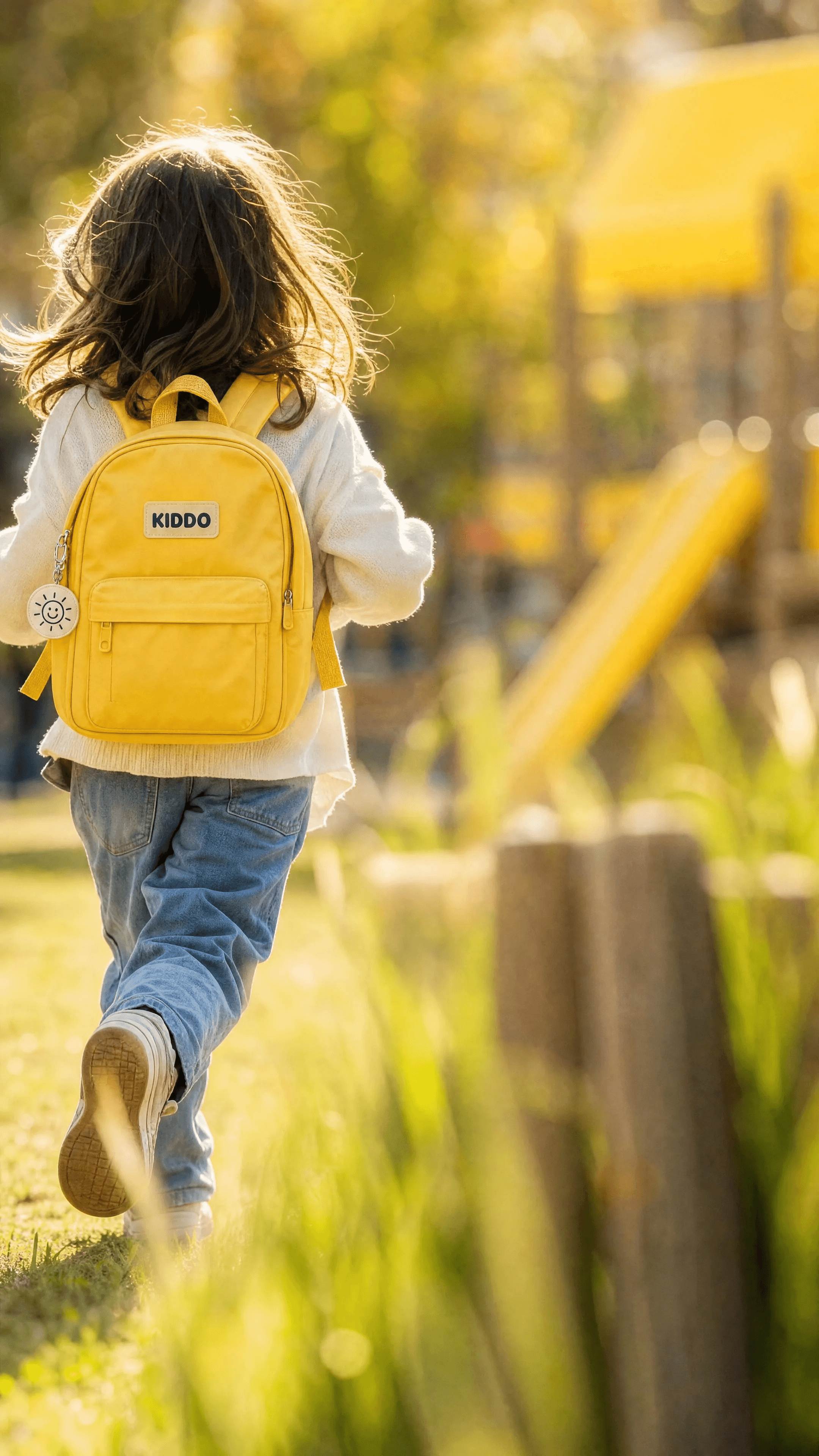 A child runs through a warm schoolyard with the butter-yellow backpack seen from behind.